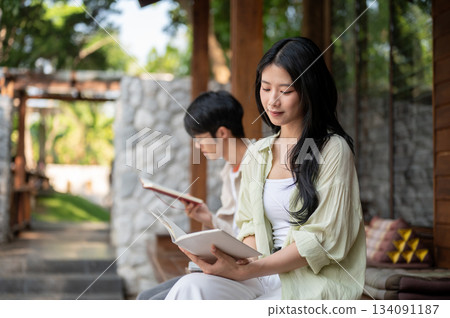 Long hair asian woman reading white book while sitting cross legged next to a man at wooden terrace. Long hair asian woman reading white book while sitting cross legged next to a man at wooden terrace. 134091187