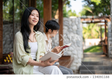 Long hair asian woman looking away from a book in hand while sitting next to a man at wooden terrace Long hair asian woman looking away from a book in hand while sitting next to a man at wooden terrace 134091188
