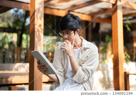 Asian man wearing glasses look at tablet with hand touching chin sitting in garden wooden corridor. Asian man wearing glasses look at tablet with hand touching chin sitting in garden wooden corridor. 134091194