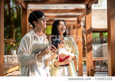 Asian woman and male friend standing looking ahead while holding laptop and book in wooden corridor. 134091203