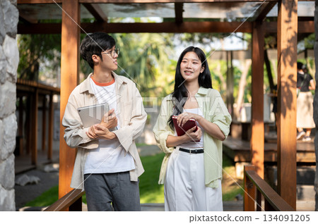 Asian friends or a man and a woman holding books and standing talking together in wooden corridor. 134091205