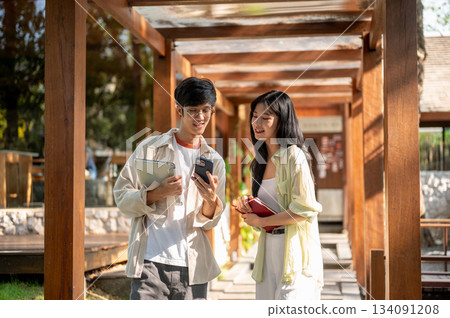 Smiling asian woman and male friend standing looking at phone with happy face in a wooden corridor. Smiling asian woman and male friend standing looking at phone with happy face in a wooden corridor. 134091208