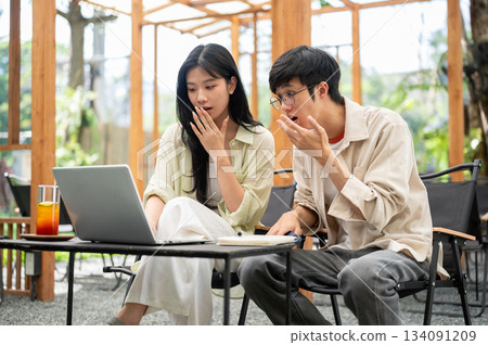 Asian woman and male friend looking at laptop screen with surprised or shocking face in garden cafe Asian woman and male friend looking at laptop screen with surprised or shocking face in garden cafe 134091209