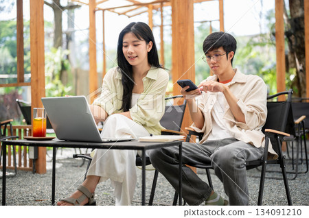 Asian woman smiling and pointing at laptop while male friend holding a phone at table in garden cafe 134091210