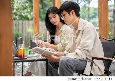Asian man with glasses holding notebook and reading together with female friend in garden cafe. 134091214