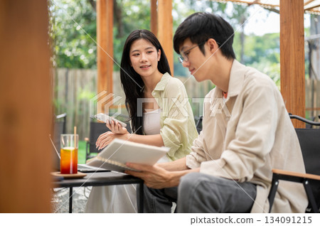 Asian man wearing glasses holding a notebook listening to female friend talking in garden cafe. 134091215