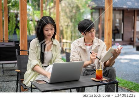 Asian woman looking at laptop sitting with a man wearing glasses reading a notebook at folding table Asian woman looking at laptop sitting with a man wearing glasses reading a notebook at folding table 134091222