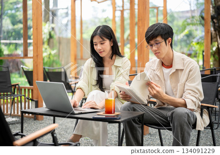 Asian woman looking and typing on laptop while a man reading a notebook sit at folding table in cafe Asian woman looking and typing on laptop while a man reading a notebook sit at folding table in cafe 134091229