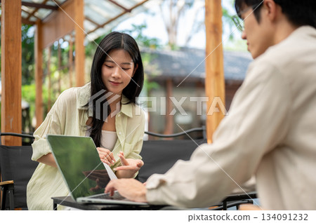 Smiling asian woman leaning to look at friend's laptop screen while sitting on folding chair in cafe 134091232