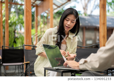 Asian woman leaning to look at laptop screen with her friend while sitting on folding chair in cafe. 134091233