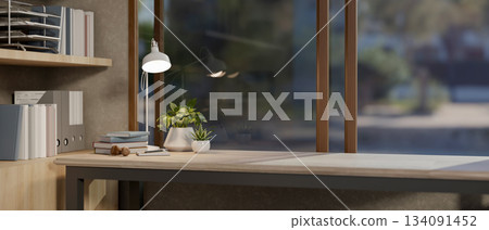 Books and potted plants next to a lamp on wooden table under windows in a living room or bedroom. 134091452