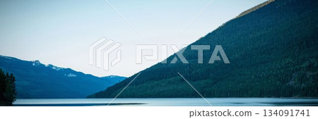 Depicting flat design lake reflecting steep forested mountain slope and snow peaks under clear sky Depicting flat design lake reflecting steep forested mountain slope and snow peaks under clear sky 134091741