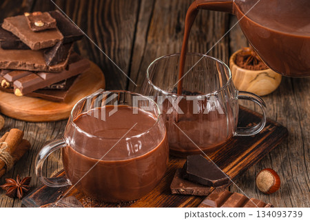 Pouring hot chocolate into glass cups with dark chocolate pieces and spices on rustic wooden table, cozy winter drink concept Pouring hot chocolate into glass cups with dark chocolate pieces and spices on rustic wooden table, cozy winter drink concept 134093739