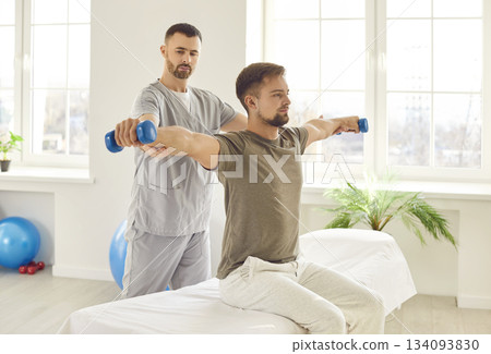 Man patient sitting on the couch in rehabilitation clinic doing sport exercise using dumbbells with nurse. 134093830