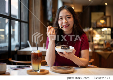 Young asian woman smiling and holding spoon enjoy eating chocolate cake aside orange coffee in cafe. Young asian woman smiling and holding spoon enjoy eating chocolate cake aside orange coffee in cafe. 134095474