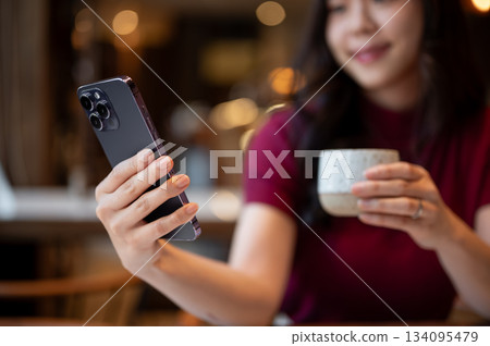 Close up of blurred asian woman holding tiramisu cup and looking at smartphone while sitting in cafe 134095479