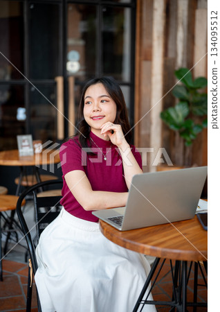 Asian woman putting hand under chin looking away from laptop while sitting at wooden table in cafe Asian woman putting hand under chin looking away from laptop while sitting at wooden table in cafe 134095512
