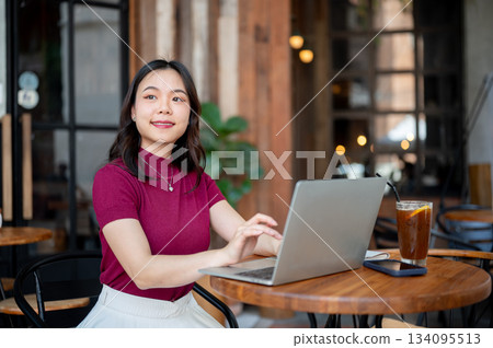 Asian woman looking away from work on laptop while sitting on metal chair at wooden table in cafe. Asian woman looking away from work on laptop while sitting on metal chair at wooden table in cafe. 134095513