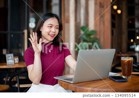 Asian woman looking and waving at laptop having video call or online class at wooden table in cafe. Asian woman looking and waving at laptop having video call or online class at wooden table in cafe. 134095514