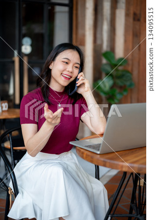 Smiling asian woman talking on phone and looking at laptop while sitting at wooden table in a cafe. Smiling asian woman talking on phone and looking at laptop while sitting at wooden table in a cafe. 134095515