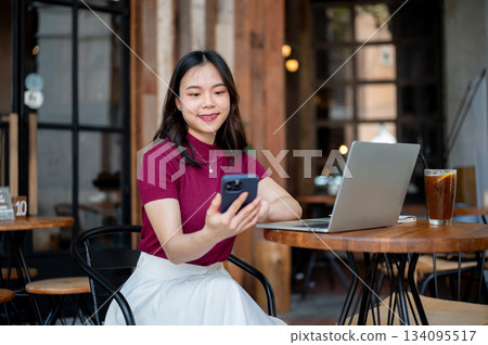Asian woman holding and looking at phone aside laptop and coffee sitting at wooden table in cafe. Asian woman holding and looking at phone aside laptop and coffee sitting at wooden table in cafe. 134095517