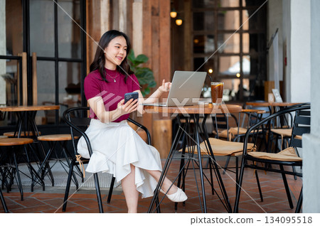 Asian woman holding phone aside and looking at laptop aside coffee sitting at wooden table in cafe. Asian woman holding phone aside and looking at laptop aside coffee sitting at wooden table in cafe. 134095518