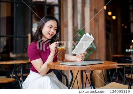 Asian woman holding straw drinking coffee and reading book aside laptop sit at wooden table in cafe. Asian woman holding straw drinking coffee and reading book aside laptop sit at wooden table in cafe. 134095519