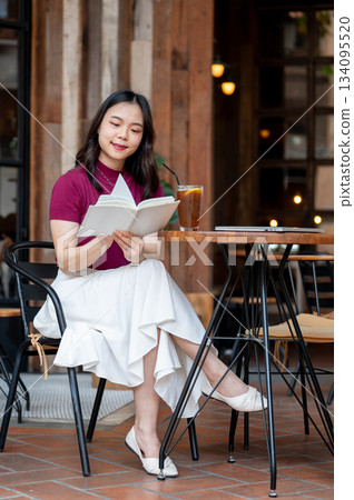Asian woman reading a book aside coffee and laptop sitting cross legged at wooden table in cafe. Asian woman reading a book aside coffee and laptop sitting cross legged at wooden table in cafe. 134095520