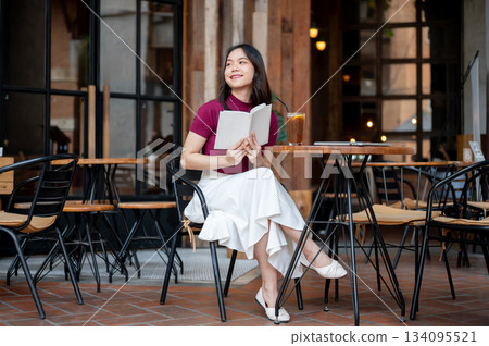 Asian woman holding book aside coffee and laptop while sitting cross legged at wooden table in cafe. Asian woman holding book aside coffee and laptop while sitting cross legged at wooden table in cafe. 134095521