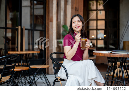 Asian woman holding straw drinking coffee while sitting cross legged at wooden table in cafe. Asian woman holding straw drinking coffee while sitting cross legged at wooden table in cafe. 134095522
