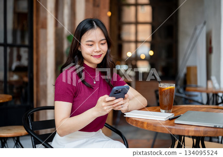 Asian woman holding and looking at smartphone aside coffee while sitting at wooden table in a cafe. Asian woman holding and looking at smartphone aside coffee while sitting at wooden table in a cafe. 134095524