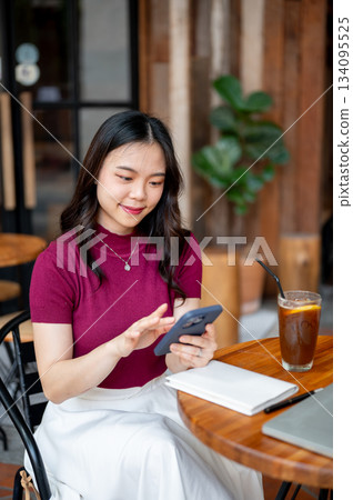 Asian woman holding looking at phone over book aside coffee while sitting at wooden table in a cafe. Asian woman holding looking at phone over book aside coffee while sitting at wooden table in a cafe. 134095525