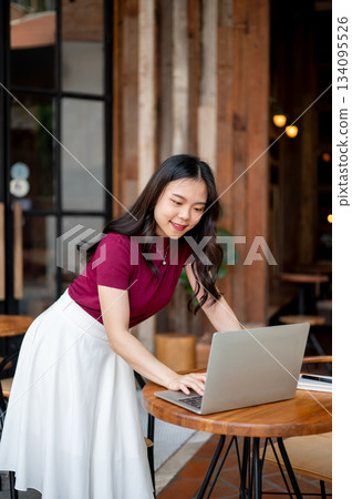 Asian young woman standing and leaning to looking at laptop on wooden table in an open air cafe. Asian young woman standing and leaning to looking at laptop on wooden table in an open air cafe. 134095526