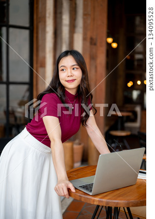 Pretty asian woman standing and leaning to looking at laptop on wooden table in open air cafe Pretty asian woman standing and leaning to looking at laptop on wooden table in open air cafe 134095528