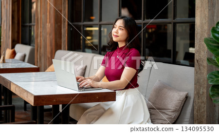 Pretty asian woman typing and looking away laptop on marble table sitting at sofa in open air cafe Pretty asian woman typing and looking away laptop on marble table sitting at sofa in open air cafe 134095530