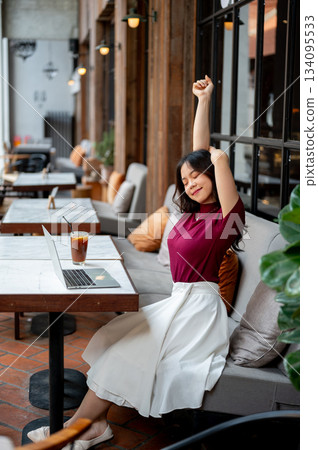 Pretty asian woman closing eyes stretching arms in front of laptop on table sitting on sofa in cafe. Pretty asian woman closing eyes stretching arms in front of laptop on table sitting on sofa in cafe. 134095533