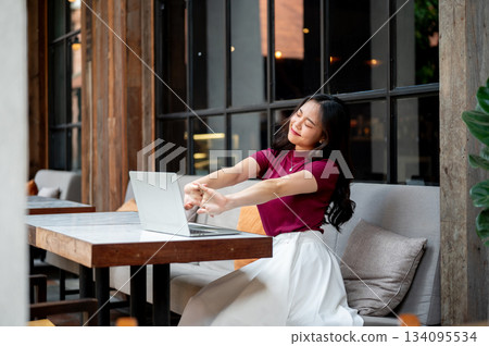 Happy asian woman closing eyes stretching arms in front of laptop on table sitting on sofa in cafe. 134095534