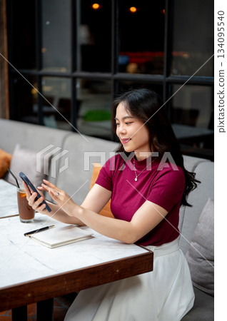 Smiling asian woman looking and touching on phone aside coffee as sitting on sofa at table in cafe 134095540
