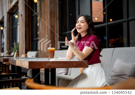 Asian woman talking on phone with open palm aside coffee on table while sitting on sofa in the cafe. Asian woman talking on phone with open palm aside coffee on table while sitting on sofa in the cafe. 134095542
