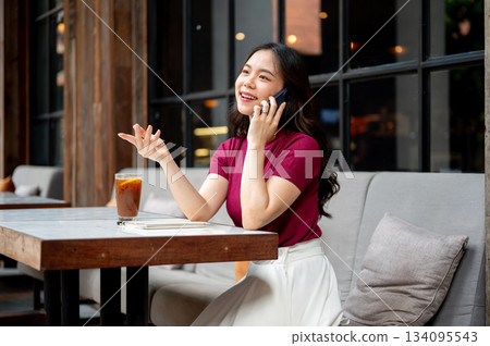 Asian woman talking on phone hand explaining aside coffee on table while sitting on sofa in the cafe 134095543