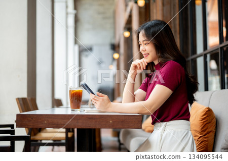 Asian woman holding and looking at phone aside coffee on table while sitting on sofa in the cafe. Asian woman holding and looking at phone aside coffee on table while sitting on sofa in the cafe. 134095544