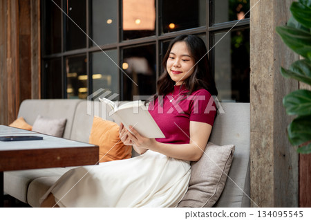 Smiling asian woman holding and reading a book while sitting leaning against sofa in the cafe. 134095545
