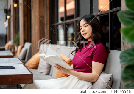 Pretty asian woman holding and reading a book while sitting leaning against sofa in the cafe. Pretty asian woman holding and reading a book while sitting leaning against sofa in the cafe. 134095546