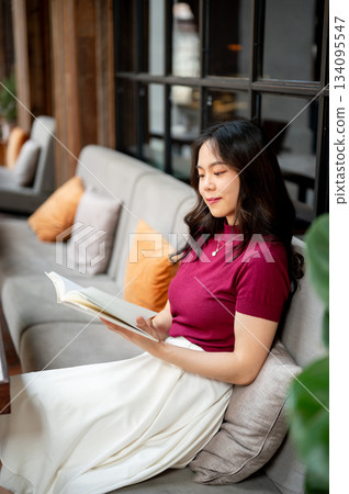 Young asian woman holding and reading a book while sitting leaning against sofa in the cafe. 134095547