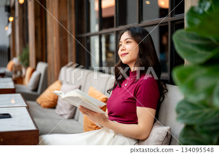 Young asian woman holding a book and looking away while sitting leaning against sofa in the cafe. Young asian woman holding a book and looking away while sitting leaning against sofa in the cafe. 134095548