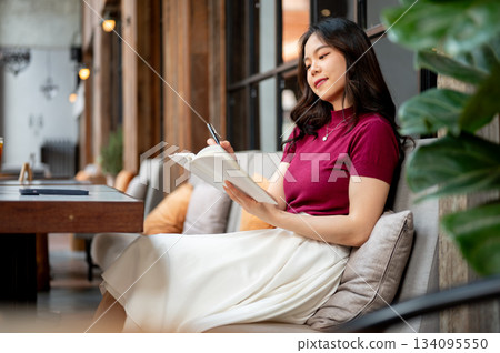 Pretty asian woman holding pen and looking at notebook while sitting leaning against sofa in a cafe. Pretty asian woman holding pen and looking at notebook while sitting leaning against sofa in a cafe. 134095550