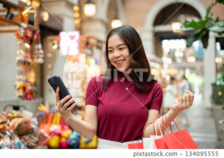 Asian woman holding shopping bags looking at phone standing beside souvenir cart in a market or mall Asian woman holding shopping bags looking at phone standing beside souvenir cart in a market or mall 134095555
