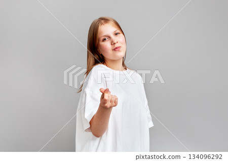 Girl posing in bright studio, making trendy finger heart gesture, love and positivity. Soft lighting, clean neutral background, modern youth culture vibe, playful expression 134096292