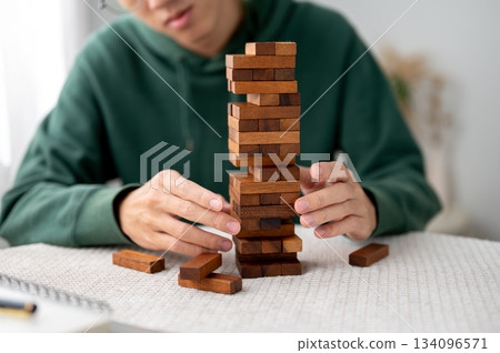 A man looking and hand taking out a piece of wooden block in tower building game while playing alone A man looking and hand taking out a piece of wooden block in tower building game while playing alone 134096571
