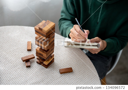 Close up a man holding pen writing in notebook aside a wooden blocks of tower building game on table 134096572